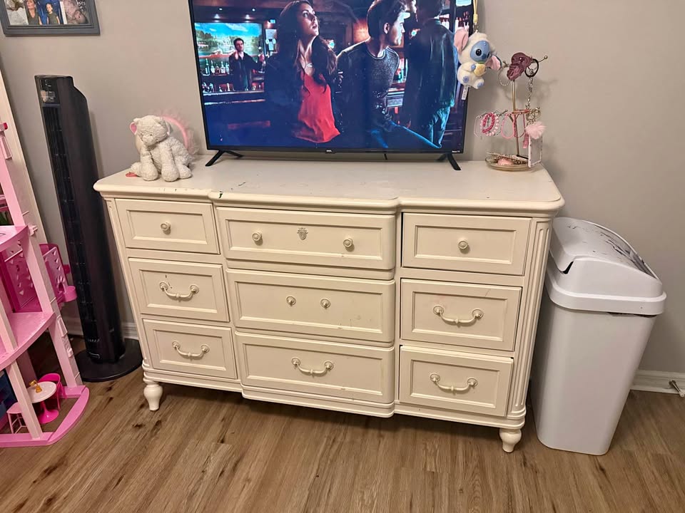 A white worn-out dresser with a television displaying a show, next to a pink shelf and a trash can.